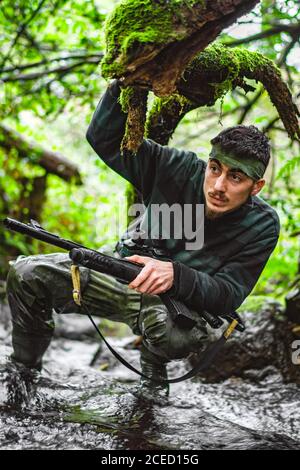 Soldier or revolutionary member or hunter in camouflage in the river observing the gun in his hand Stock Photo