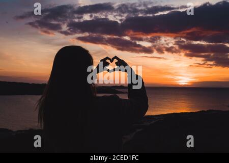 Back view of Woman standing at the ocean in sunset lights and gesturing heart with fingers. Stock Photo