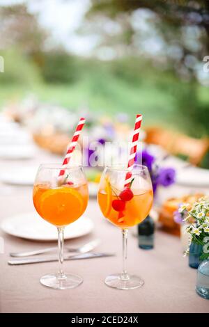 Glasses of delicious Aperol spritz drink with straw placed on banquet table with appetizers and fresh flowers in garden Stock Photo