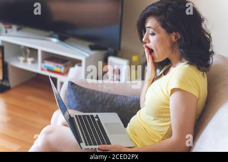 Surprised woman having online video chat with friends via laptop covering her mouth with hand, surprised expression, Woman can't believe talking with Stock Photo