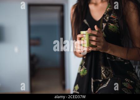 Side view beautiful passionate brunette Hispanic Woman in dress looking away and holding mug of hot in flat Stock Photo
