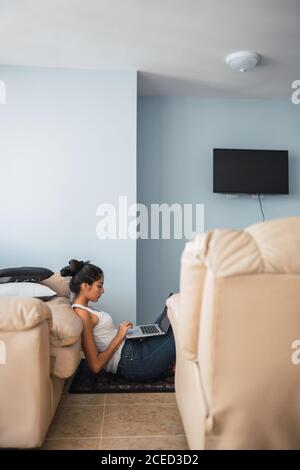 Beautiful brunette Hispanic Woman typing on laptop and sitting on floor on little carpet near settee and window Stock Photo