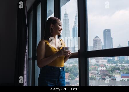 Smiling pretty Asian Woman standing with cup at the window and looking away. Stock Photo