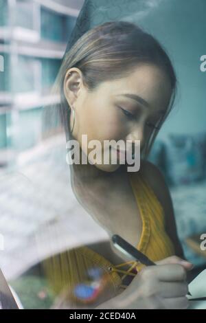 View through window of pretty Asian Woman standing and taking notes at home. Stock Photo