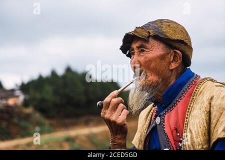 DONGCHUAN RED, YUNNAN, CHINA - JUNE 14, 2018: Relaxed senior male in bright traditional clothes sitting on ground smoking wooden tobacco pipe and petting dog sitting?nearby looking away on blurred background of Dongchuan red lands in Chinese province of Yunnan Stock Photo