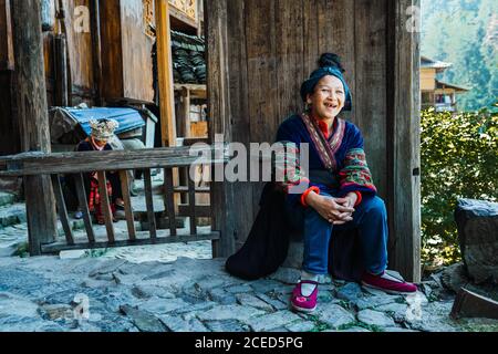 GUINZHOU, CHINA - JUNE 14, 2018: Happy female of Miao ethnic group of Chinese Guizhou province sitting in bright traditional clothes on terrace with old wooden railing and walls with weathered village buildings and trees around on sunny summer day Stock Photo