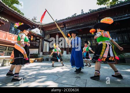 GUINZHOU, CHINA - JUNE 14, 2018: Miao ethnic group man in blue clothes playing traditional musical instrument and women in bright local costumes dancing around him during festival in village in Guizhou province of China Stock Photo