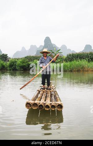 Smiling Asian elderly man in straw hat approaching standing on bamboo raft with paddle in hands amidst Qu?y S?n river with high karst mountains and grassy bank on background on cloudy day in Chinese province of Guangxi Stock Photo