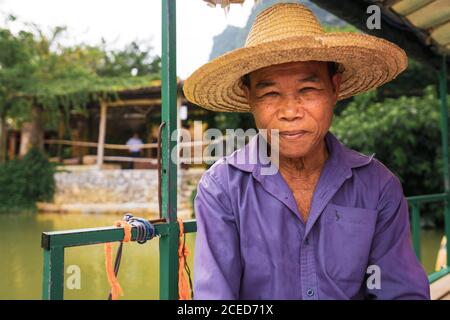 Smiling aged Asian fisherman in straw hat looking at camera sitting at Quây Son river harbor in small village surrounded by trees and mountains in Guangxi province of China Stock Photo