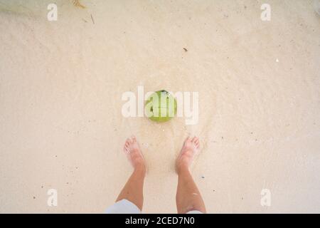 Crop shot from above of barefoot man standing on white sand of tropical beach with green coconut in water below, China Stock Photo