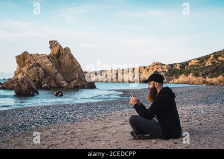Side view of cheerful bearded man sitting on the beach and using smartphone. Stock Photo