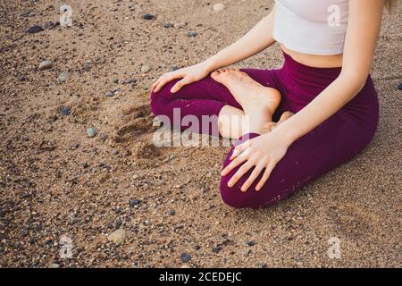 Crop unrecognizable Woman sitting on wet sand and meditating. Stock Photo