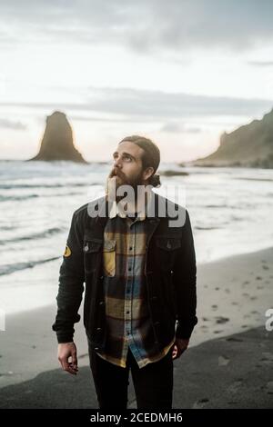 Stylish bearded guy looking up white standing on sandy shore near amazing waving sea Stock Photo