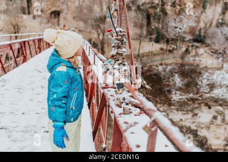 Side view of cute child in warm clothes standing on modern bridge and looking at bunch of love padlocks on winter day in countryside Stock Photo