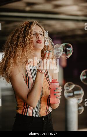 Content stylish curly blonde in summer outfit blowing soap bubbles underground Stock Photo