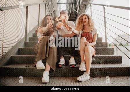 Three cheerful friends laughing and burning sparkles while sitting on metal stairs on city street Stock Photo