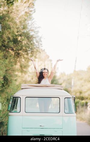 Pretty young lady smiling and keeping hands up while standing in hatch of retro van during trip in nature Stock Photo