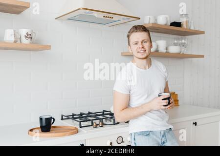 Portrait young man smiles drink fresh coffee in ceramic black mug breakfast. Bright kitchen background Stock Photo