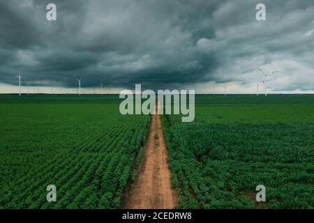 Countryside dirt road through agricultural fields with wind turbines in background on dark cloudy overcast afternoon
