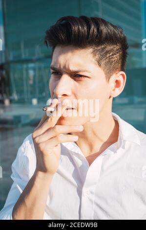 Young handsome with modern hairstyle wearing white shirt and smoking cigarette on street in sunlight Stock Photo