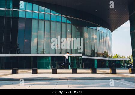 Side view at distance of modern man jumping on blocks on pavement against modern shiny skyscraper Stock Photo