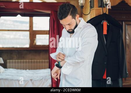 Young handsome man with dark hair and beard putting on white shirt in bedroom with new black costume hanging on clothes hanger Stock Photo
