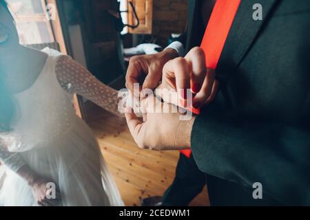 Crop view of man in black costume helping young bride in wedding gown put on bracelet on hand on background of room Stock Photo