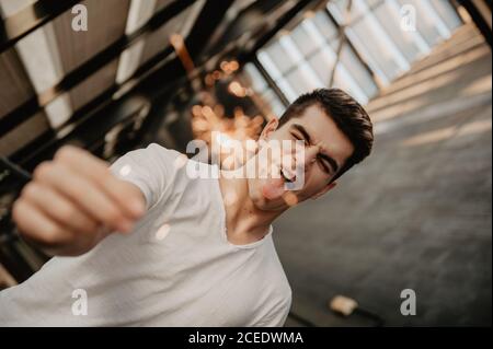 Young handsome man in white T-shirt standing inside building and holding burning Bengal light in hand Stock Photo