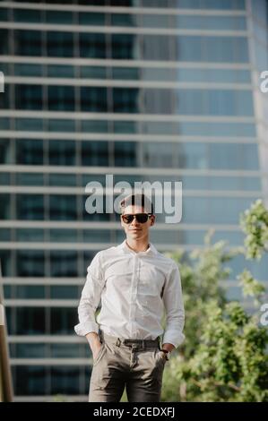 Confident young man keeping hands in pockets and looking at camera while standing on background of skyscraper on sunny day on modern city street Stock Photo