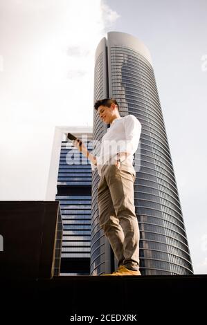 From below shot of stylish young guy smiling and using smartphone while standing near skyscraper in modern city Stock Photo