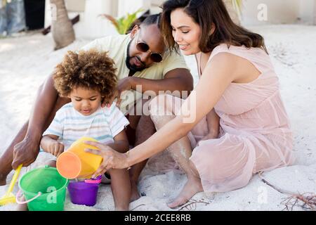 Happy African-American parents and son sitting on sand and playing with buckets in sunny day on vacation Stock Photo