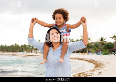 Cheerful ethnic Woman holding and carrying little son on shoulders while walking on sandy coast at seaside Stock Photo