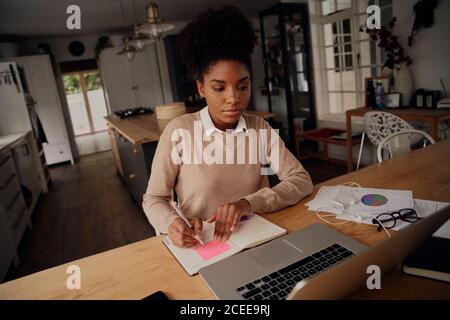 Young freelancer writing details on book while working on laptop at home during lockdown Stock Photo