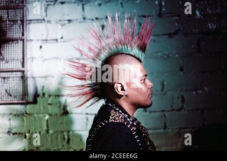 Side view of man in jacket with metal pikes wearing colorful mohawk on background of brick wall. Stock Photo