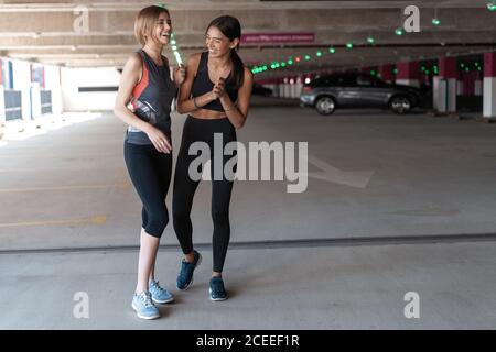Two happy smiling young athletic woman walking outdoors together after sport training, talking and hugging each other. Stock Photo