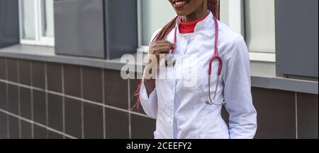 black young woman doctor, in a white coat, with a phonendoscope, holds a folder for medical papers in a rue, against the background of a hospital Stock Photo