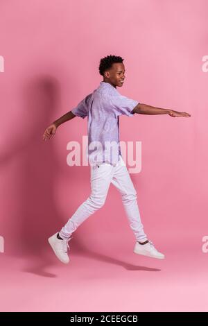 Side view of smiling young black man in white denim and shirt jumping up against pink background Stock Photo