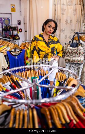 Beautiful young lady in stylish outfit leaning on clothes rail and looking at camera while standing in small store Stock Photo