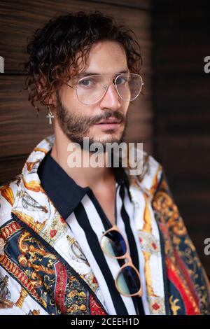 Handsome young guy with curly hair wearing trendy clothes and looking at camera while standing on blurred background of wall Stock Photo