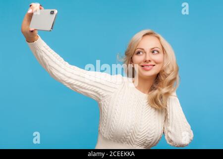 Trendy beautiful woman in knitted sweater posing holding smartphone in hand, female blogger making selfie for social network. Indoor studio shot isola Stock Photo