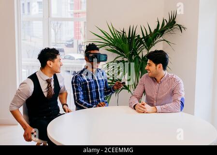 Cheerful people having fun and sitting with VR helmet at table in office. Stock Photo