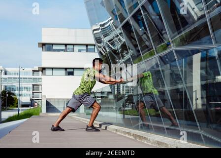 Side view of adult ethnic man stretching at glass handrail while working out on modern city street Stock Photo