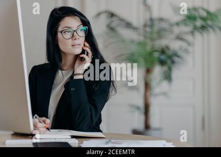 Elegant young woman in formal outfit answers phone call, writes down necessary notes in notepad, looks away with thoughtful expression, works at moder Stock Photo