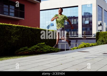 Smiling handsome ethnic man jumping while running on city street in sunny day Stock Photo