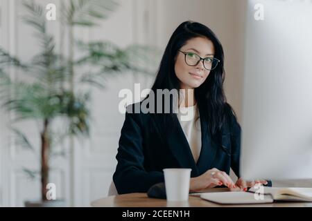 Serious successful female office worker in formal outfit, wears eyeglasses, busy with online work, poses at desktop, works on computer, reads business Stock Photo