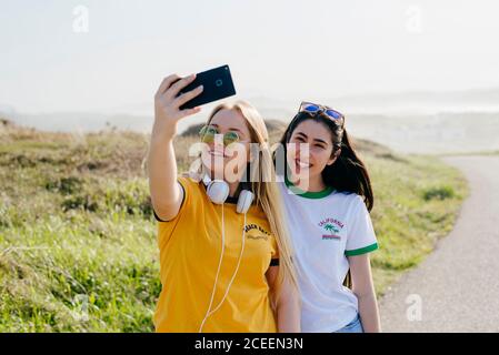 Young pretty brunette and blonde in casual outfits taking selfie with phone on background of shoreline. Stock Photo