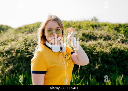 Casual young girl wearing sunglasses with yellow t-shirt and headphones on neck smiling at camera in green nature. Stock Photo
