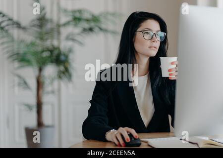 Busy young female administrative poses at desktop, drinks hot coffee from paper cup, dressed formally, develops business strategy, plans startup, invo