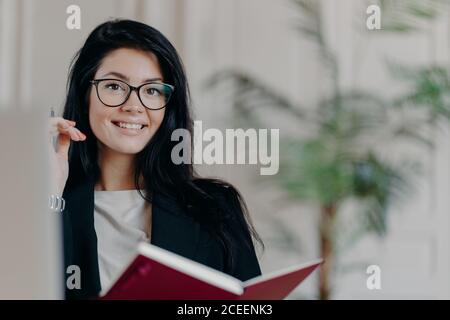 Horizontal shot of satisfied brunette freelancer works remotely from home, holds notepad and pen, makes notes, spends free time for research, fills ou Stock Photo