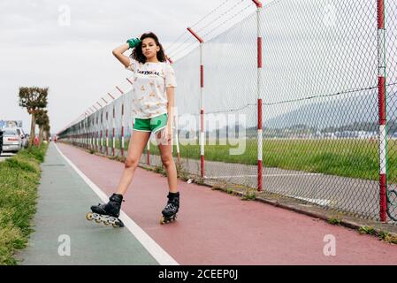 Casual teen girl in shorts and roller skates standing on lane of sports ground looking at camera. Stock Photo